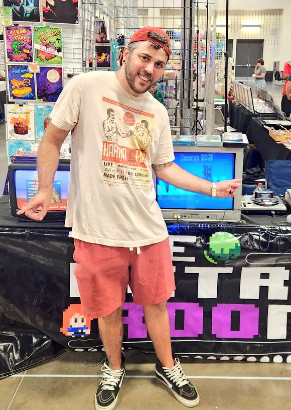A photo of John Roo standing at an exhibition stand for The Retro Room.